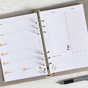 Peut inclure: Un agenda hebdomadaire blanc avec des anneaux dorés. L'agenda comporte des sections pour "mois", "notes", "à faire", "suivi des habitudes" et un suivi des habitudes quotidien. L'agenda est décoré d'un motif floral.