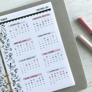 May include: A planner page with a habit tracker for November 2027. The page includes sections for exercise, healthy eating, meditation, and other habits. Pink and gray markers rest on the side. The planner is open on a light wood surface.