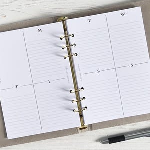 May include: A white weekly planner with a gold binder. The planner has a section for each day of the week, with lines for writing tasks and appointments. The planner also has a section for notes and a section for to-do lists.