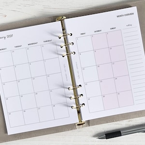 May include: Open planner showing a January 2027 calendar page. The calendar is in a light grey binder with gold rings. A pen rests on the bottom right. The calendar has a month overview section with space for notes.