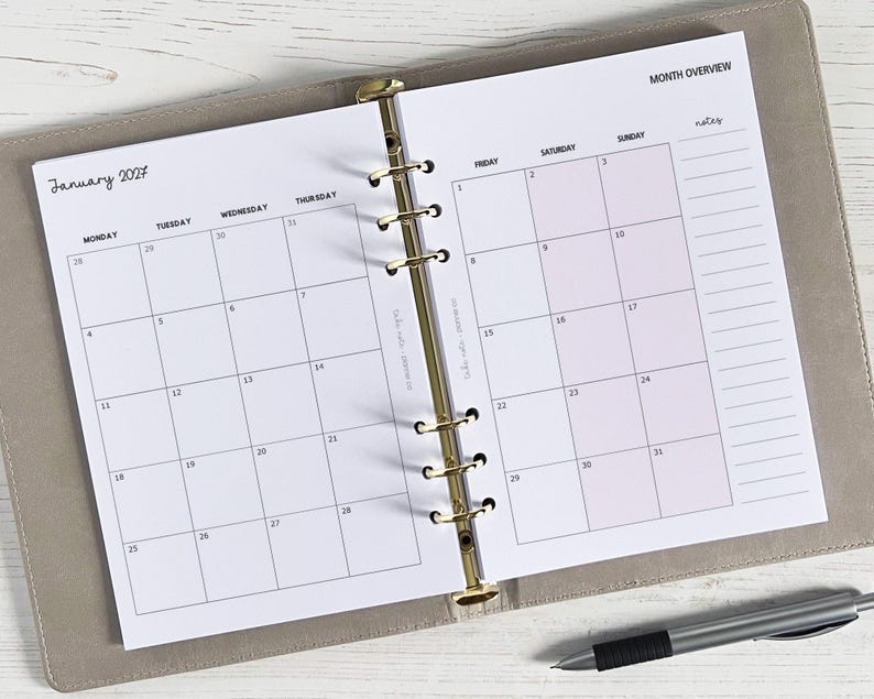 May include: Open planner showing a January 2027 calendar page. The calendar is in a light grey binder with gold rings. A pen rests on the bottom right. The calendar has a month overview section with space for notes.