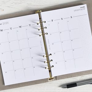 Op de afbeelding: Een witte maandplanner met een gouden ringband. De planner heeft een maandoverzicht voor januari 2026. De planner is open voor de maand januari en de dagen van de week zijn gelabeld met M, D, W, D, V, Z, Z.