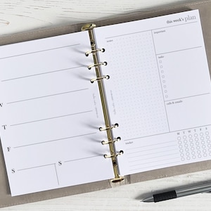 Könnte beinhalten: Ein weißer Wochenplaner mit einem goldenen Binder. Der Planer hat Abschnitte für "Diese Woche planen", "Monat", "Notizen", "Wichtig", "Aufgaben", "Anrufe & E-Mails" und "Tracker".