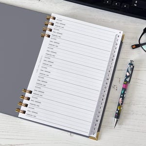 May include: A gray address book with gold spiral binding and labeled tabs. The pages are lined and include prompts for account, email, and password. A patterned pen and black glasses rest nearby on a white wooden surface.