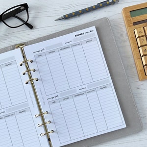 May include: A grey and white savings tracker with a gold binder ring. The tracker has columns for date, deposits, withdrawals, interest paid, and balance. The tracker is on a white wooden surface with a calculator and glasses.