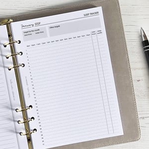 May include: A planner open to a sleep tracker page for January 2027. The page has a grid for tracking sleep hours and quality, with sections for bed and wake times. A black pen rests on the right side of the planner, which is open on a light-colored wooden surface.