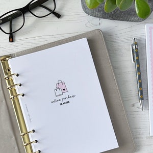 Peut inclure: Un agenda blanc avec un anneau de reliure doré. L'agenda a une page avec une illustration rose et noire de sacs de shopping et le texte "online purchase tracker".