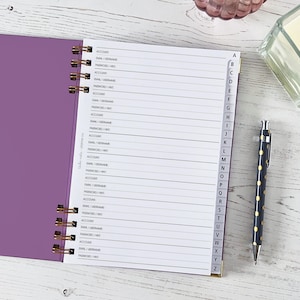 Könnte beinhalten: Ein offenes, lila Spiralnotizbuch mit linierten Seiten und Buchstaben-Reitern. Ein schwarzer Stift mit goldenen Punkten liegt neben dem Notizbuch. Die Seiten des Notizbuchs sind mit "EMAIL/USERNAME" und "PASSWORD/HINT" beschriftet.