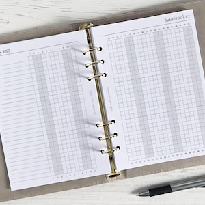 Può includere: Agenda aperto con un tracker delle abitudini per gennaio 2027. Le pagine sono bianche con un layout a griglia per il monitoraggio delle abitudini quotidiane. L'agenda è in un raccoglitore grigio chiaro con anelli dorati. Una penna d'argento poggia sulla superficie.