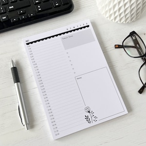 May include: A white daily planner with a black and white floral design. The planner has a column for the date, a column for the time, and a column for notes. The planner is on a white wooden surface.