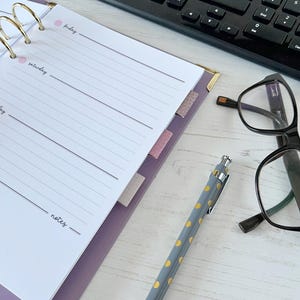 May include: A lilac planner with gold rings, lined pages, and pink tabs. A black keyboard, spectacles, and a pen with yellow spots are also visible. The planner is open to a page with the days of the week.