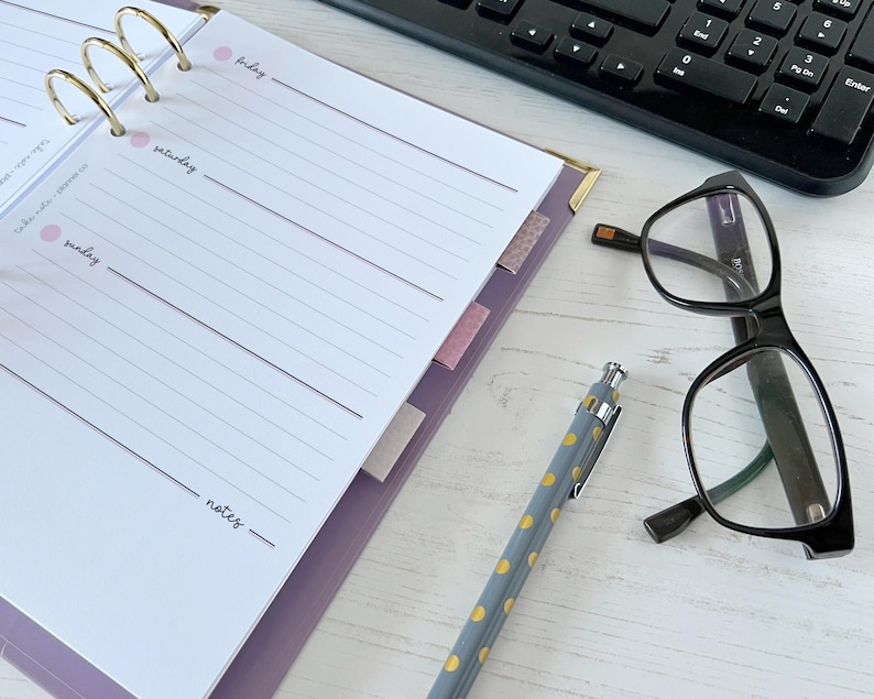 May include: A lilac planner with gold rings, lined pages, and pink tabs. A black keyboard, spectacles, and a pen with yellow spots are also visible. The planner is open to a page with the days of the week.