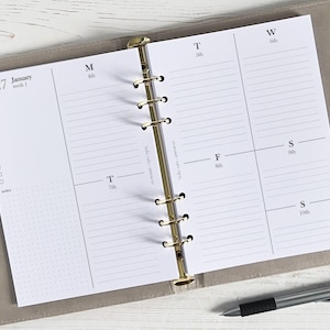 Peut inclure: Agenda hebdomadaire ouvert sur une page blanche avec des lignes noires et les dates de janvier 2027. Le planificateur est maintenu dans un classeur gris clair avec des anneaux dorés. Un stylo argenté est posé en bas à droite.