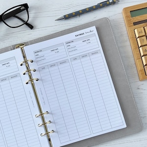 May include: A grey and white savings tracker with a gold binder. The tracker has columns for date, deposit, and balance. The tracker is on a white wooden surface with a calculator.