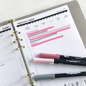 Könnte beinhalten: Eine Planerseite mit einem Produktivitätstracker für Januar 2023. Die Seite hat einen Zusammenfassungsbereich und ein Diagramm mit rosa und grauen Balken. Drei farbige Stifte, rosa, hellrosa und grau, liegen auf der Seite.