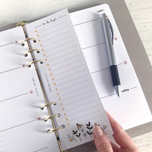 May include: A white planner with gold rings and a to-do list section. The to-do list section has a dotted line pattern and a floral design at the bottom. The planner is open to a weekly spread with the word "Week 1" at the top.