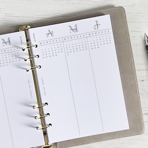 Puede incluir: Una agenda con una cubierta gris claro, abierta en una página de calendario. La página muestra los meses de abril, mayo y junio, con un diseño semanal. Un bolígrafo negro descansa en el lado derecho. La agenda está abierta sobre una superficie de madera clara.