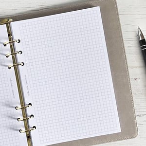 Może przedstawiać: Szary notes skórzany z złotymi kółkami i stroną w kratkę. Notes jest otwarty na pustej stronie. Czarny długopis leży na białym tle obok notesu.