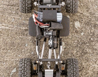 Op de afbeelding: Bovenaanzicht van een zwart en zilver RC auto chassis met vier grote, getextureerde banden. Het chassis heeft verschillende mechanische componenten, waaronder een motor, vering en elektronische onderdelen. De banden zijn zwart en hebben een robuust profiel.