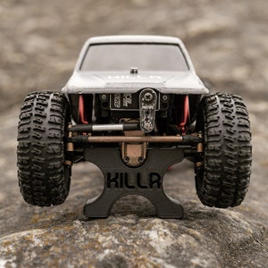 May include: A black plastic stand with the word "KILLA" printed on it. A gray and black toy truck is sitting on the stand. The truck has black tires and a red suspension.