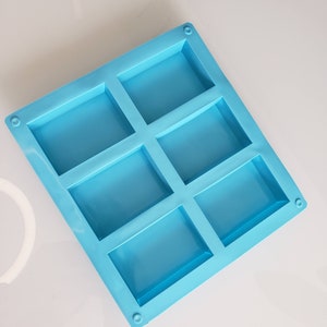 Puede incluir: Un molde de silicona azul claro con seis cavidades rectangulares. El molde está diseñado para hacer barras de jabón u otras pequeñas artesanías.