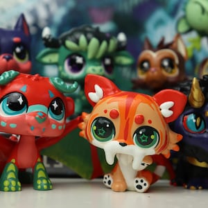 May include: A collection of small, stylized animal figurines featuring large, expressive eyes. The group includes a red dragon with green wings, an orange and white saber-toothed cat, a purple creature with gold horns, and various other colorful fantasy pets.