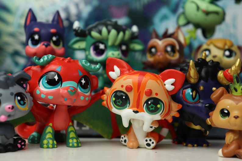 May include: A collection of small, stylized animal figurines featuring large, expressive eyes. The group includes a red dragon with green wings, an orange and white saber-toothed cat, a purple creature with gold horns, and various other colorful fantasy pets.