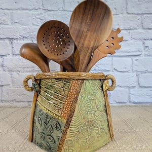 May include: A ceramic utensil crock with a textured, multi-paneled surface featuring embossed leaf, mushroom, and geometric patterns in shades of green and brown, holding a set of dark brown wooden kitchen spoons and a pasta server.