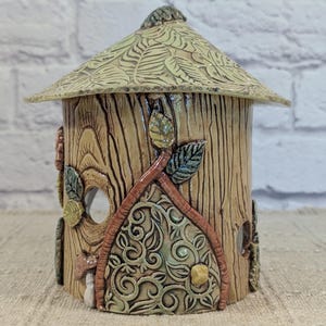 May include: A cylindrical ceramic lantern with a conical roof, featuring a textured wood grain finish. The structure includes a decorative arched door with intricate scrollwork, applied leaf accents in green and brown, and circular cutouts.