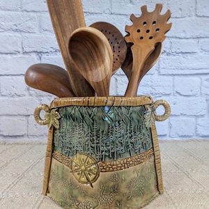 May include: A ceramic utensil crock with a forest and mountain relief design, featuring a compass and pinecone motif, holding a set of five wooden kitchen tools including a spatula, spoons, and a pasta server with a natural wood grain.