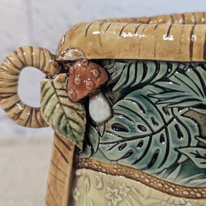 May include: A ceramic vessel featuring a textured tan rim and a handle with a coiled design. The exterior displays a relief of a brown mushroom, green leaves, and a lower section with a cream-colored floral pattern and green foliage.
