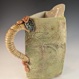 May include: A light green ceramic pitcher featuring an embossed forest floor pattern with various mushrooms and ferns. The handle is textured to resemble a tree branch and is adorned with small sculpted mushrooms and a single dark leaf.
