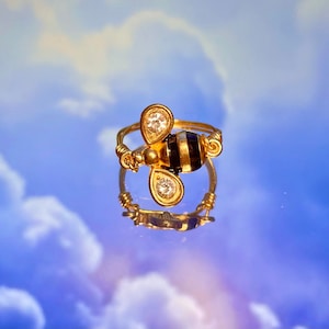 May include: A gold ring with a black and yellow bee charm with clear rhinestones. The ring is on a blue and white background.