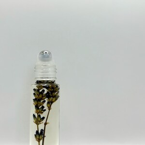 May include: A clear glass roll-on bottle filled with a clear liquid and sprigs of lavender. The bottle has a silver rollerball and cap. The lavender sprigs are a dark brown colour. The background is plain white.
