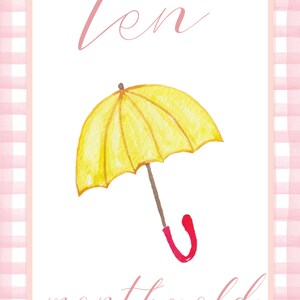 May include: A pink and white gingham border surrounds a white card with the text "ten months old" written in pink cursive. A yellow umbrella with a red handle is illustrated in the center of the card.