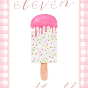 May include: A white popsicle with pink frosting and colorful sprinkles on a wooden stick. The text "eleven months old" is written in pink cursive.