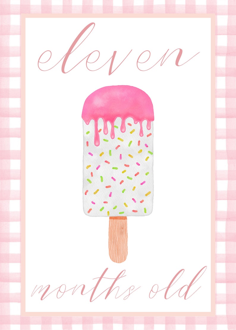 May include: A white popsicle with pink frosting and colorful sprinkles on a wooden stick. The text "eleven months old" is written in pink cursive.