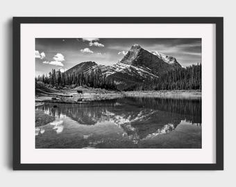 May include: Black and white framed print of a mountain landscape. The image features a lake reflecting the mountains and trees. A small animal stands near the lake's edge. The sky has clouds. The print is in a black frame.