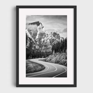 Pode incluir: Uma impressão artística emoldurada em preto e branco apresentando uma estrada de asfalto sinuosa que leva a um pico de montanha imponente e escarpado, parcialmente obscurecido por nuvens, com uma densa floresta de pinheiros na base.