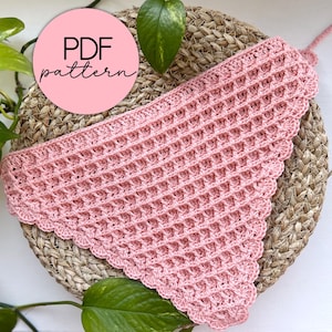 May include: A pink crocheted triangle scarf with a scalloped edge, displayed on a woven straw mat. The text "PDF pattern" is visible on a pink circle.