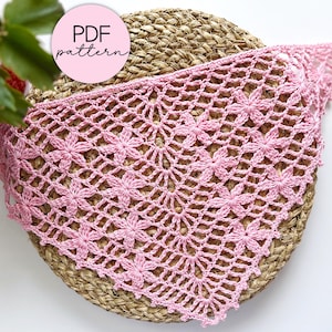 Puede incluir: Un chal de ganchillo rosa claro con un patrón floral, apoyado sobre una superficie tejida. El chal está hecho de un hilo delicado y presenta un diseño floral repetido. Un círculo rosa con el texto "PDF pattern" está en la parte superior izquierda.