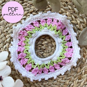 Può includere: Un elastico per capelli all'uncinetto bianco fatto a mano con un motivo floreale rosa e verde. L'accessorio per capelli ha una forma circolare con un'apertura centrale e un bordo smerlato. Il testo "PDF pattern" è in un cerchio rosa.