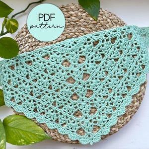 May include: A light green, crocheted shawl with a scalloped edge, resting on a woven mat. The shawl features an open, lacy design. A circular sticker with the text "PDF pattern" is visible. Green leaves and vines are in the background.