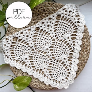 May include: A white, crocheted doily with a scalloped edge, resting on a woven, circular mat. The doily features a detailed, openwork pattern. The words "PDF pattern" are in a white circle in the upper left corner. Green leaves and vines are in the background.