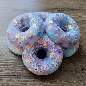 Könnte beinhalten: Vier pastellblaue, donutförmige Untersetzer aus Harz mit verschiedenen schimmernden Glitzer, Muscheln und weißen Buchstabenperlen.