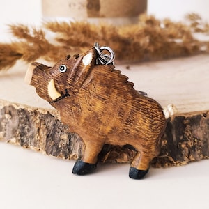 May include: A wooden wild boar keychain with a light brown finish. The boar has a white snout and black hooves. The keychain is attached to a silver ring.