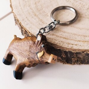 May include: A wooden boar keychain with a brown and black painted design. The keychain has a silver metal ring and chain.