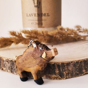 May include: A wooden wild boar figurine with a brown and black paint scheme. The figurine has a smiling face with a small tusk on one side. The figurine is sitting on a wooden surface.