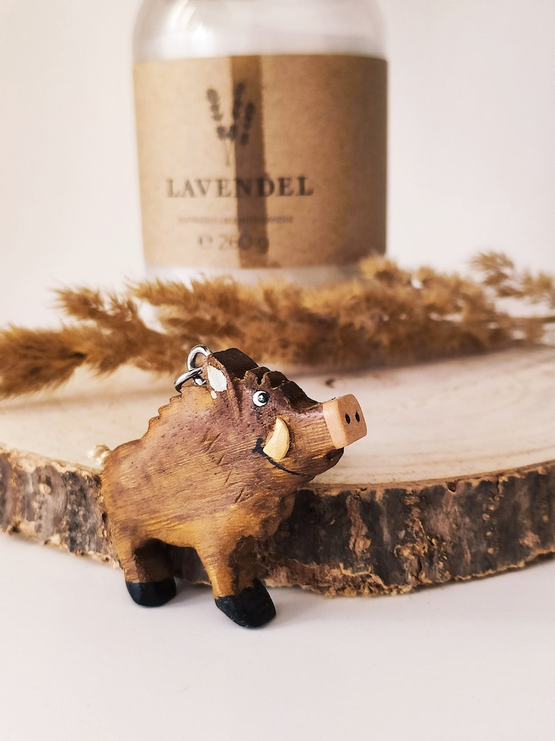 May include: A wooden wild boar figurine with a brown and black paint scheme. The figurine has a smiling face with a small tusk on one side. The figurine is sitting on a wooden surface.