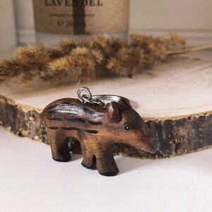 May include: Wooden keychain shaped like a wild boar with brown and black stripes. The keychain is on a wooden surface.